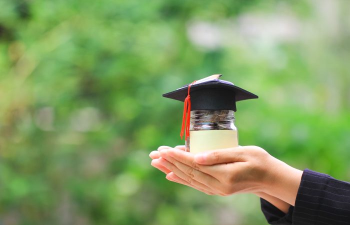 Woman hand holding coins money in glass bottle with graduates hat on natural green background, Saving money for education concept